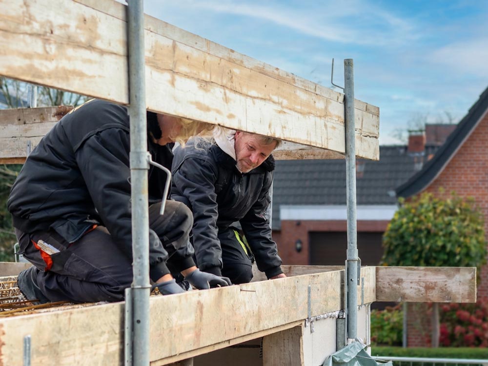 Zwei Bauarbeiter knien auf einem Gerüst und arbeiten an einer Holzkonstruktion auf einer Baustelle